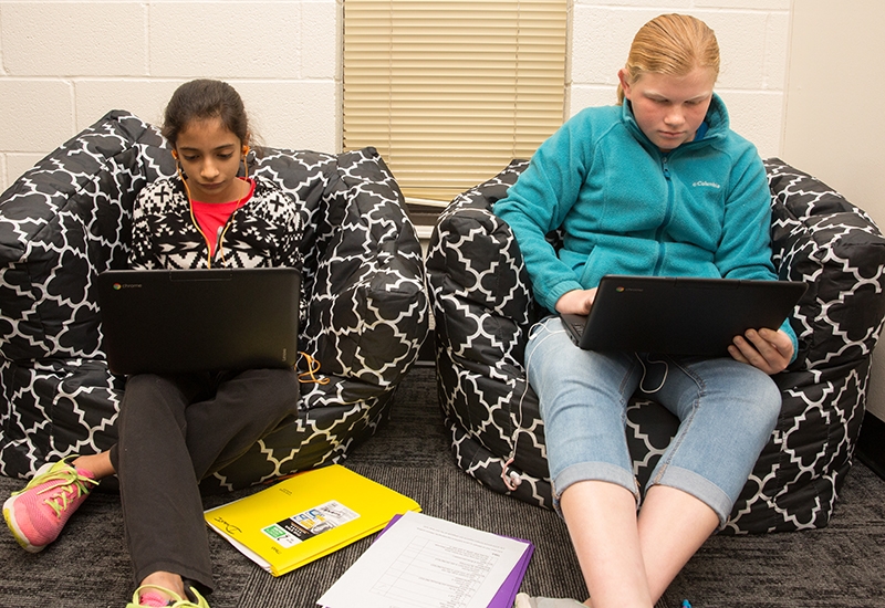 Two students sit side-by-side on beanbag chairs, looking down at laptops
