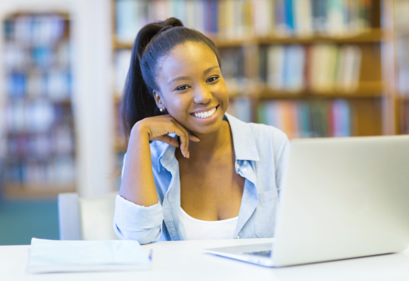 Girl sits in front of laptop inside library, smiling at camera

