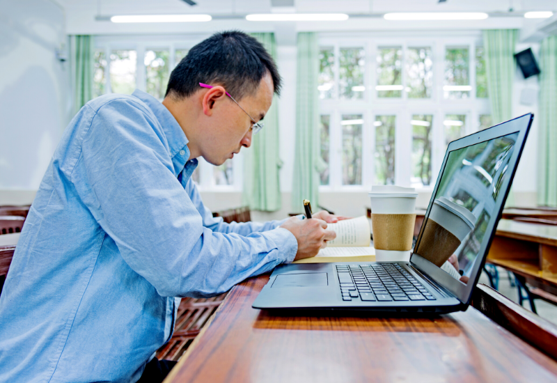 Man writing down at desk, sitting in front of laptop