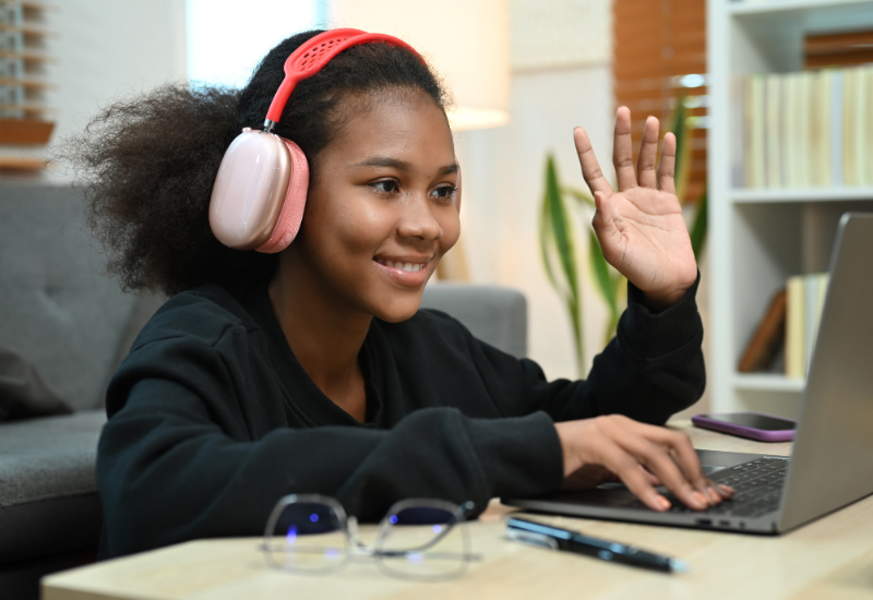Student wearing headphones waves at laptop screen, smiling
