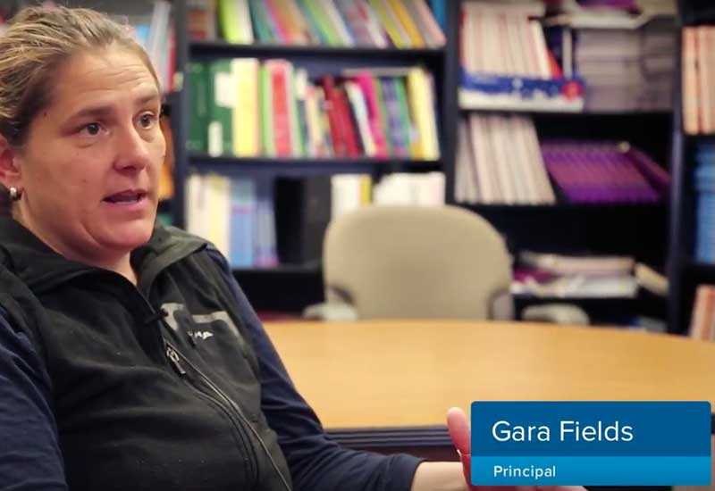 Woman speaks to camera inside library, with the label Gara Fields, principal in the corner
