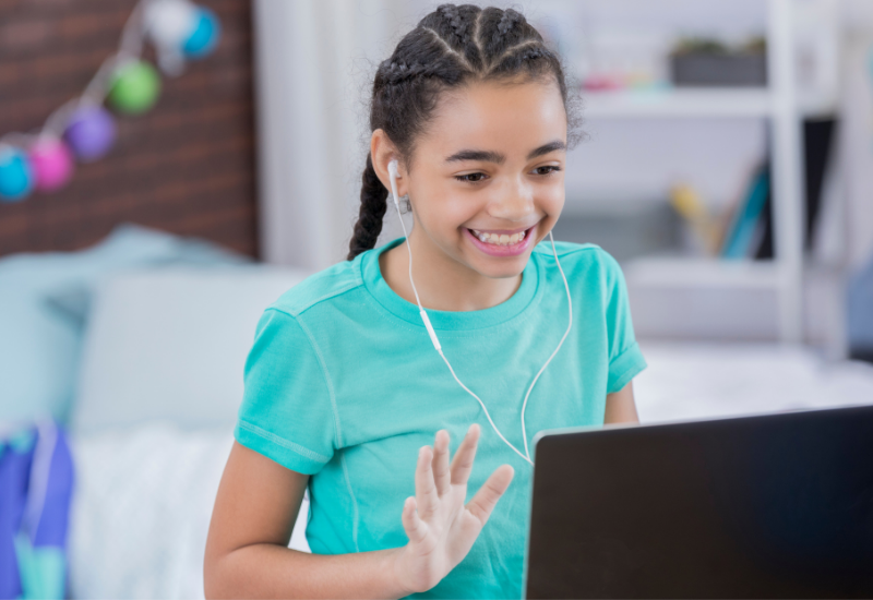 Student smiles at laptop screen, waving her hand, wearing earphones
