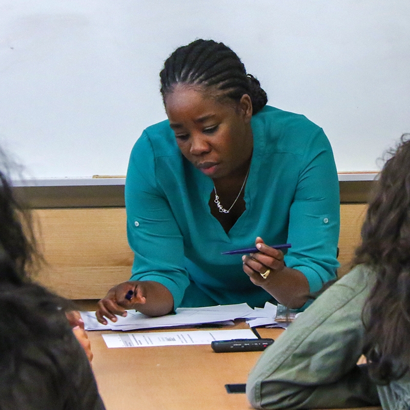 Teacher sitting at a desk with a small group of students looking at some papers with a pen in hand
