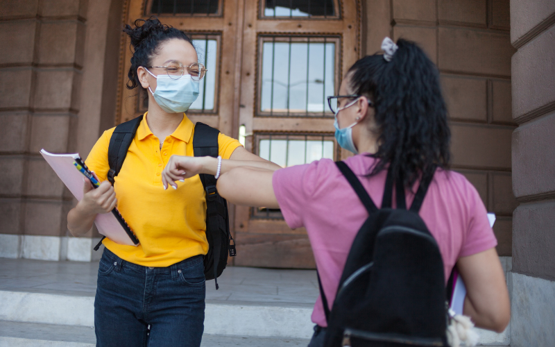 Two students wearing bookbags and face masks bump elbows in greeting
