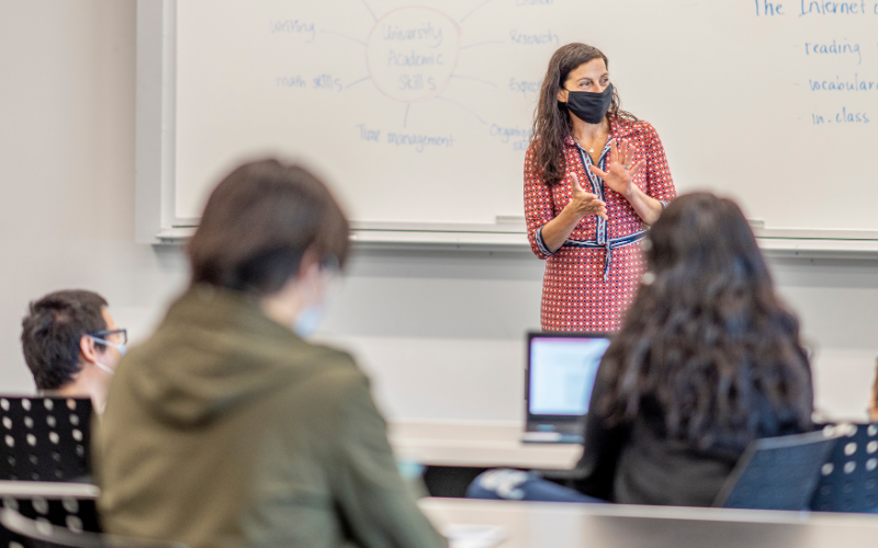 Teacher wearing mask stands in front of whiteboard in classroom as students sit scattered at desks, some with laptops in front of them, all wearing masks
