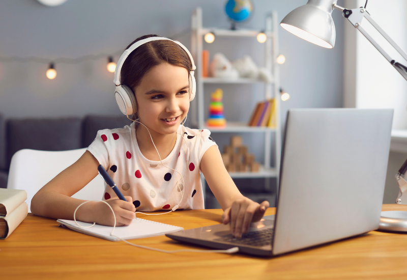 Young girl sits in bedroom, working on laptop, listening to audio over headphones
