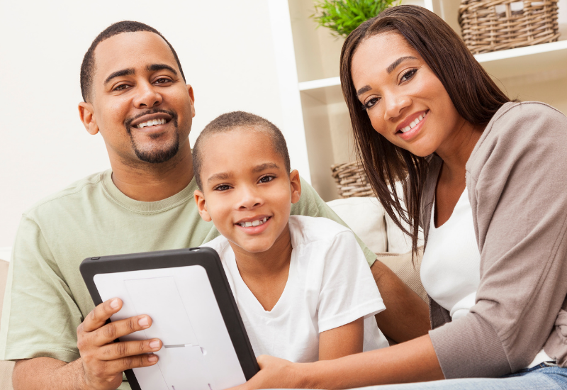 A child and his parents look at camera, smiling, while holding a tablet
