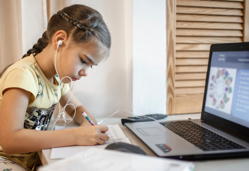 Young girl writes on paper, sitting in front of laptop, listening to audio through earbuds
