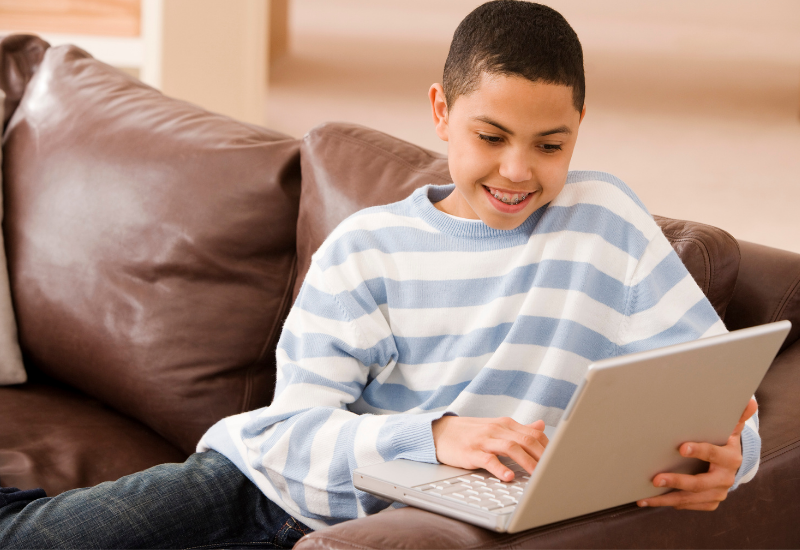 Student sits on couch, smiling down at his laptop
