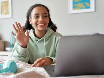 A girl sitting on a bed using a laptop, waving at screen
