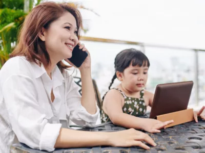 A woman sitting at a table talking on a cell phone while a child sits with tablet next to her
