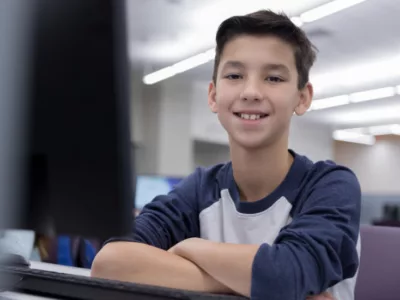 A young boy sitting at a desk with his arms crossed, smiling
