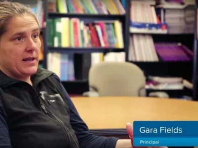 Woman speaks to camera inside library, with the label Gara Fields, principal in the corner
