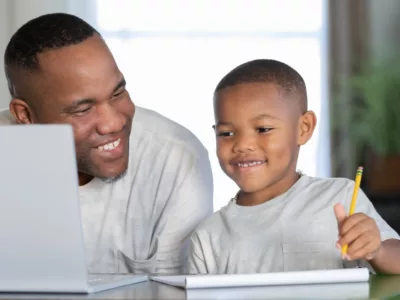 Man works with young student, smiling at laptop, pencil in hand

