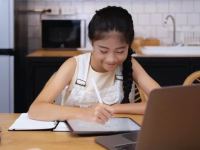 Student writes on tablet, at home in kitchen, in front of laptop
