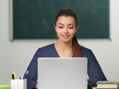 Teacher sits in classroom in front of blackboard, looking down at laptop
