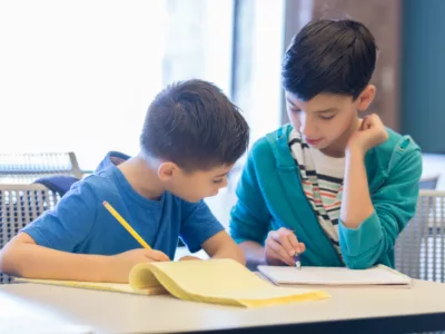 Two students sitting at desk, working together, in front of notebooks, writing
