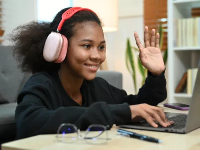 Student wearing headphones waves at laptop screen, smiling
