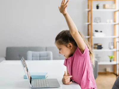 A little girl sitting at a table with a laptop, raising her hand
