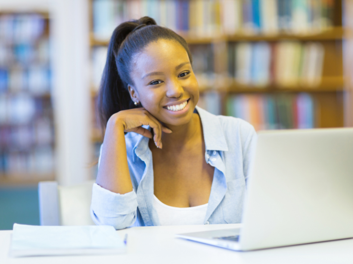 Girl sits in front of laptop inside library, smiling at camera
