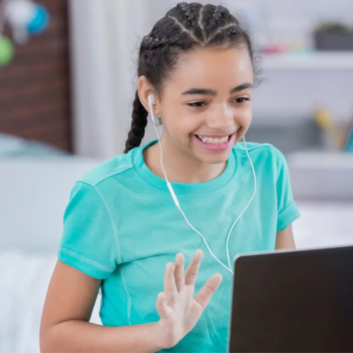 Student smiles at laptop screen, waving her hand, wearing earphones