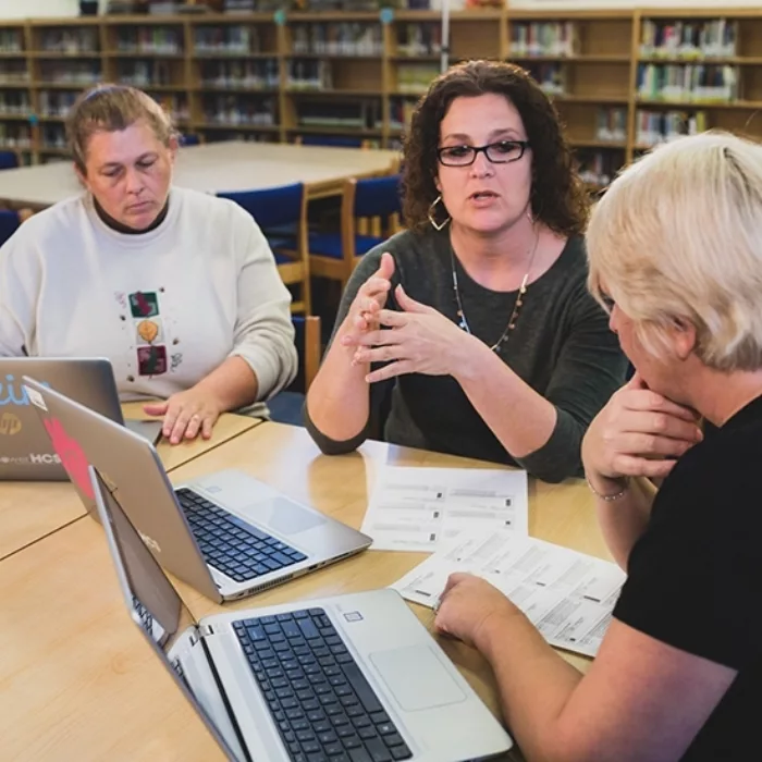 Three teachers sit at desk with laptops, speaking with each other as they work
