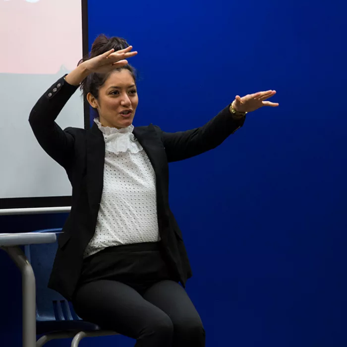 Woman in suit sits in front of white and blue background, gesturing enthusiastically
