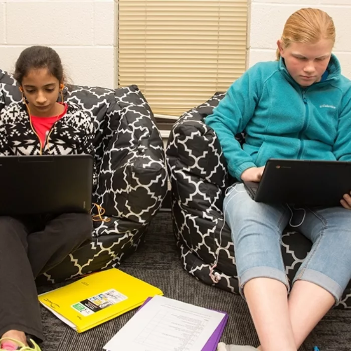 Two students sit side-by-side on beanbag chairs, looking down at laptops