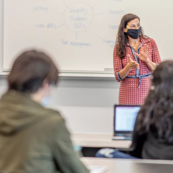 Teacher wearing mask stands in front of whiteboard in classroom as students sit scattered at desks, some with laptops in front of them, all wearing masks