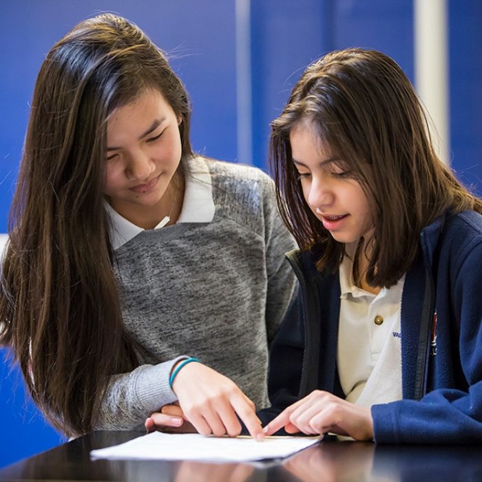 Two students look down together at a paper on desk
