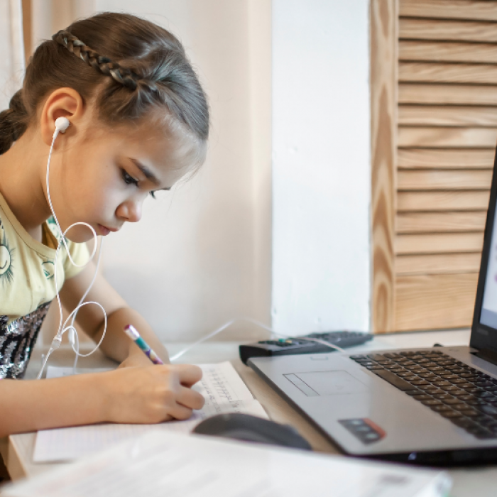Young girl writes on paper, sitting in front of laptop, listening to audio through earbuds