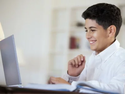 Boy sits in front of an open laptop, smiling, with one of his hands starting to make a triumphant fist
