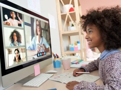 Girl sits in front of large desktop monitor, smiling at screen, where video of other students and a teacher can be seen
