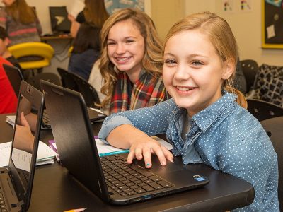 Students smile at camera while sitting at laptops
