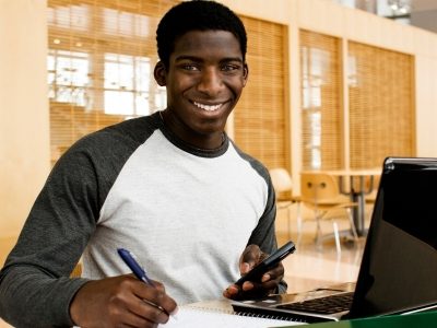 Teen sits in front of laptop, writing on paper, smiling
