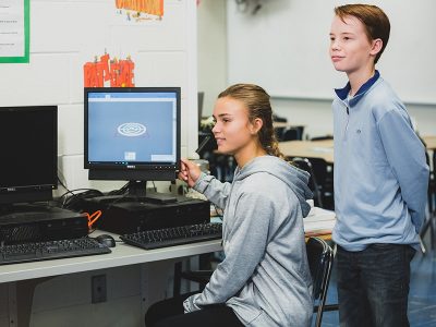 One student sits in front of computer, gesturing toward screen and smiling, while another student stands nearby, smiling
