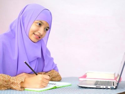 Girl writes in notebook while looking at laptop and smiling
