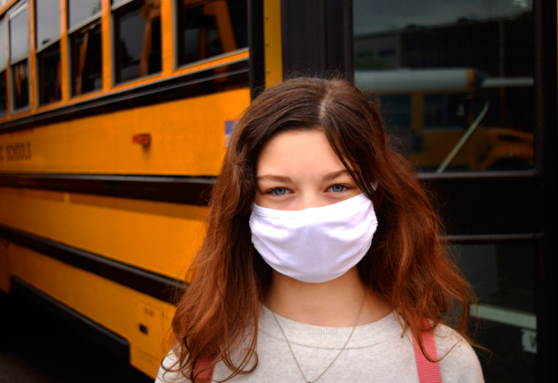 Child wearing mask, standing in front of school bus
