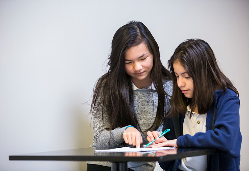 Two students working together at desk, looking on together at paper
