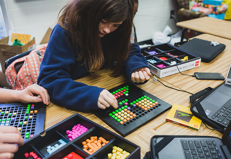 Student works on project, placing colorful blocks in a small grid
