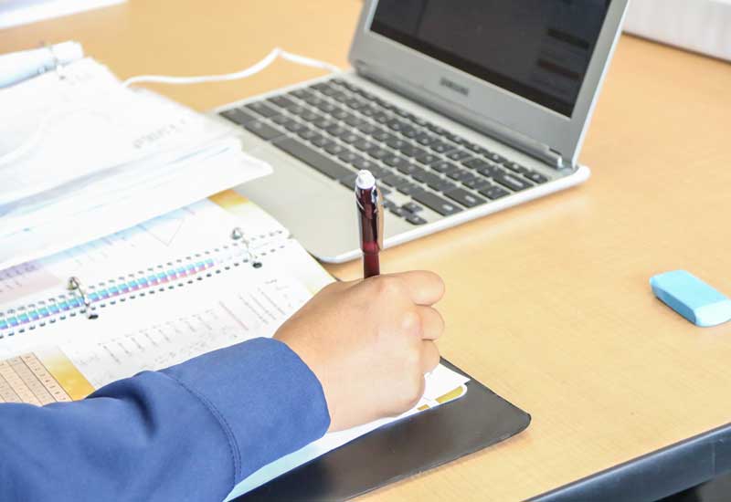 Photo of student arm writing in notebook with laptop in background