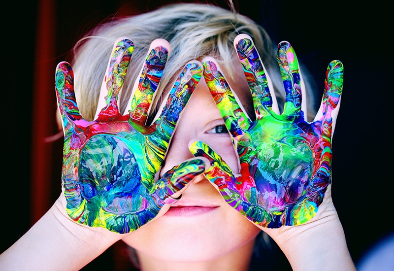 A child's raised hands with bright, multicolored swirls of paint on both palms with the child's eyes peeking through their spread fingers.
