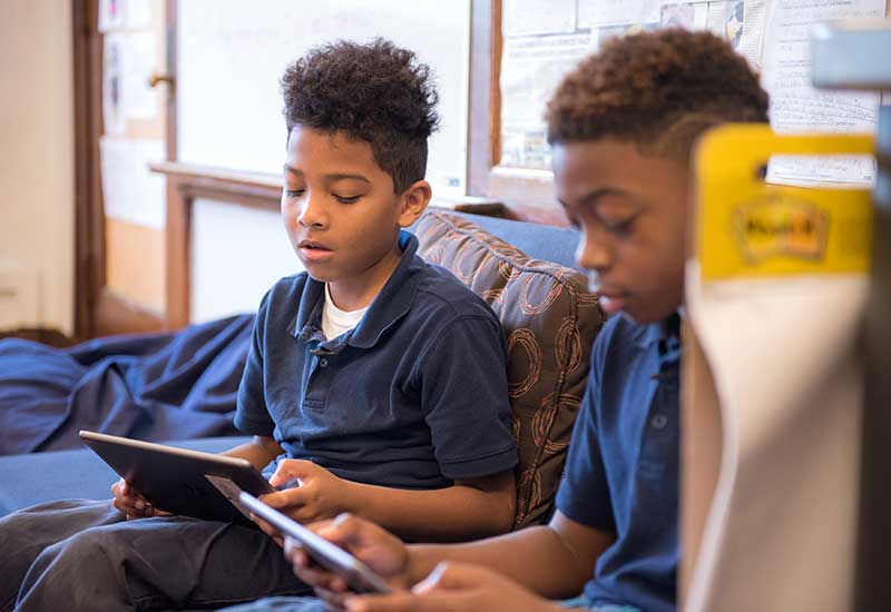 Two students sit on couch, reading from tablets
