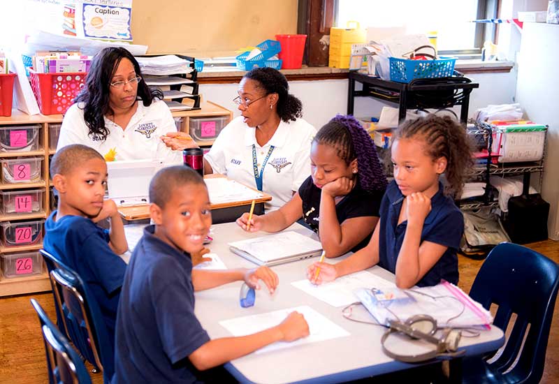 Four students sit at grouped desks alongside two educators inside classroom