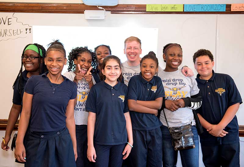 Eight students stand with teacher in front of white board, smiling
