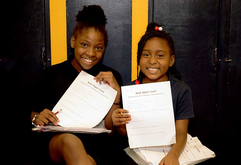 Two students sit beside each other in hallway and hold up reports, smiling
