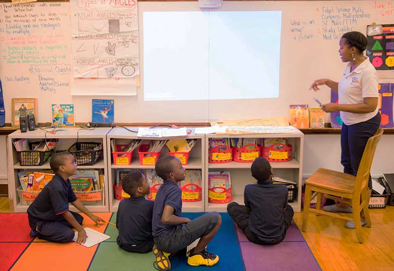 Four students sit on colorful floor, looking up at teacher as she gestures at white board