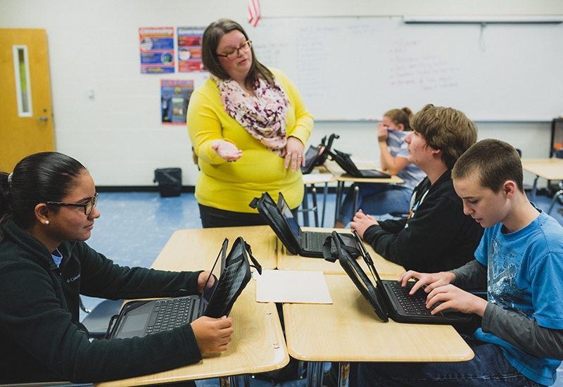 Teacher speaks with student while he sits at joined desks with other students who are looking at their laptops