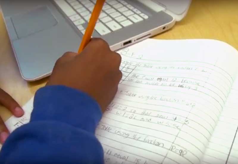 Photo closeup of student working in workbook with device closeby
