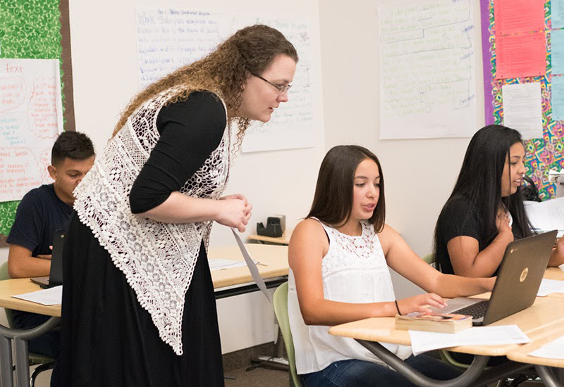 Teacher stands behind student on laptop at desk, looking over student's shoulder as she speaks
